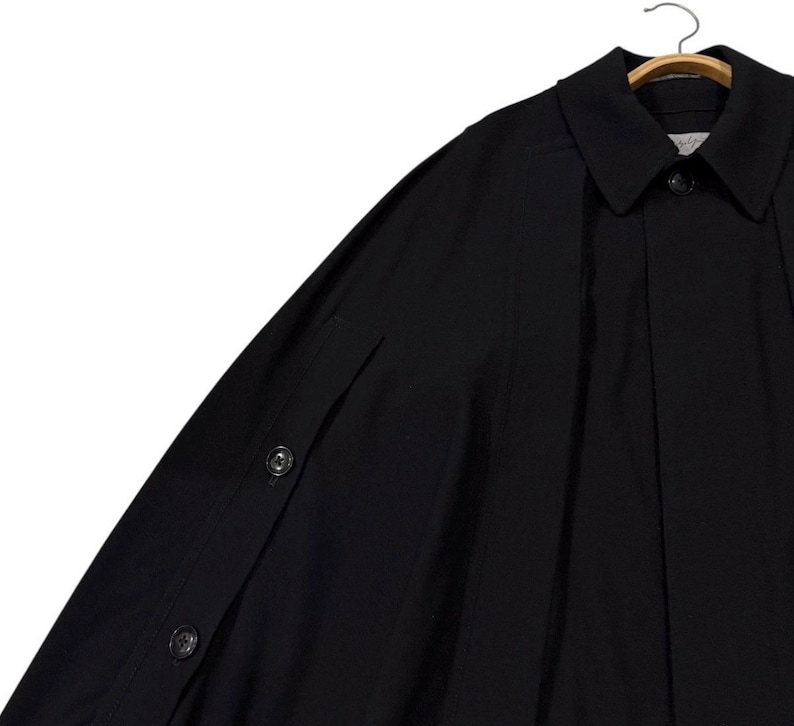 May include: A black cape-style coat with a collar and button closure. The coat is hanging on a wooden hanger. The sleeves have button details. The fabric appears to be a smooth, solid material.