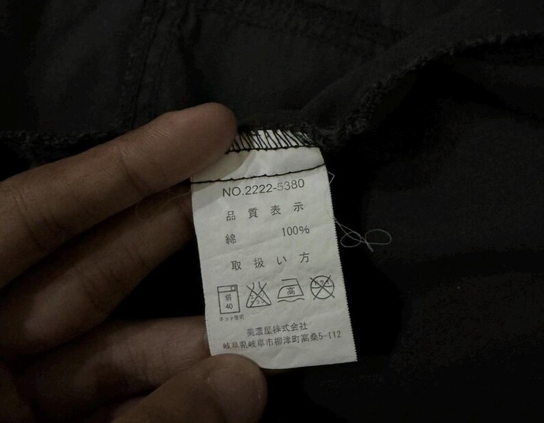Puede incluir: Primer plano de una prenda negra con una etiqueta blanca. La etiqueta muestra texto japon&eacute;s, incluyendo "NO.2222-5380", "100% algod&oacute;n" y s&iacute;mbolos de cuidado. La tela parece ligeramente arrugada.