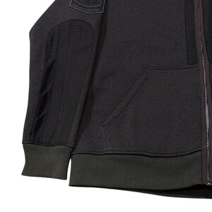 May include: Dark grey zip-up hoodie with black elbow and shoulder patches. The hoodie has a ribbed hem and cuffs, and a front pocket. The zip runs down the centre.