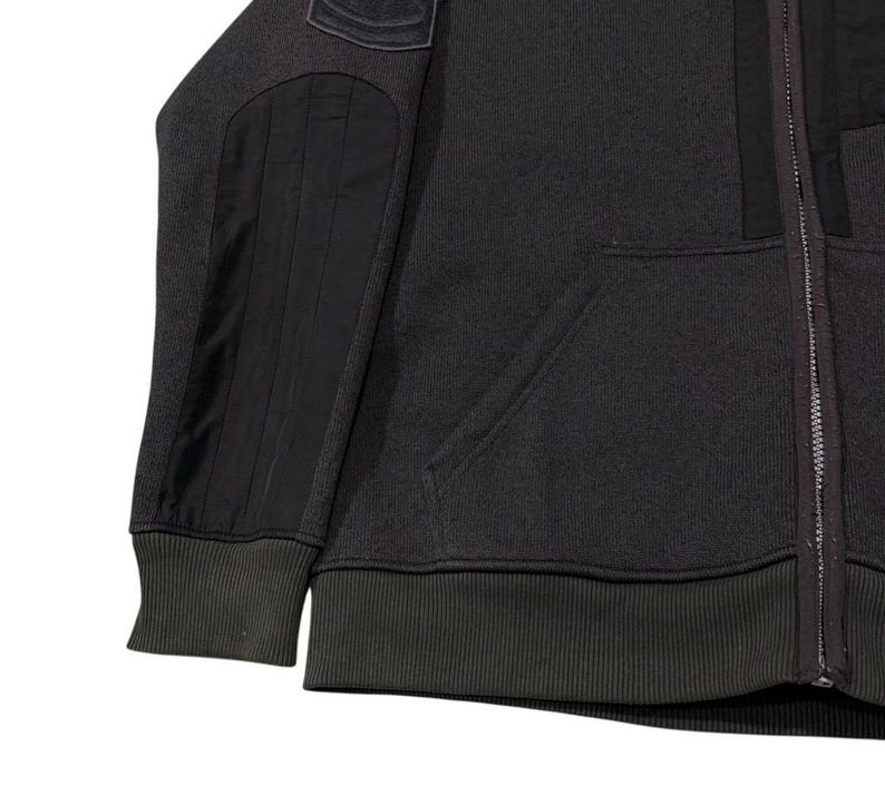 May include: Dark grey zip-up hoodie with black elbow and shoulder patches. The hoodie has a ribbed hem and cuffs, and a front pocket. The zip runs down the centre.