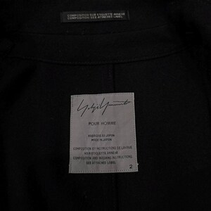 May include: Black garment interior with a label that reads "POUR HOMME" and "FABRIQUE AU JAPON MADE IN JAPAN". The label also includes text in French and English regarding composition and washing instructions. The garment has a tag that says "COMPOSITION SUR ETIQUETTE ANNEXE COMPOSITION: SEE ATTACHED LABEL".