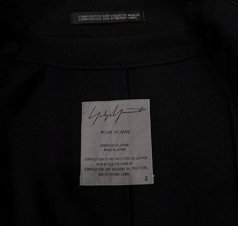 May include: Black garment interior with a label that reads "POUR HOMME" and "FABRIQUE AU JAPON MADE IN JAPAN". The label also includes text in French and English regarding composition and washing instructions. The garment has a tag that says "COMPOSITION SUR ETIQUETTE ANNEXE COMPOSITION: SEE ATTACHED LABEL".
