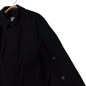 May include: Black coat with a classic collar and button details. The coat is hanging on a wooden hanger. The coat has a buttoned sleeve detail.