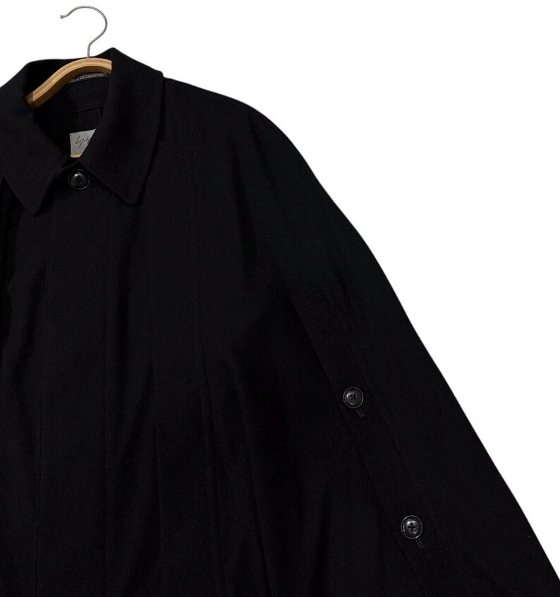 May include: Black coat with a classic collar and button details. The coat is hanging on a wooden hanger. The coat has a buttoned sleeve detail.
