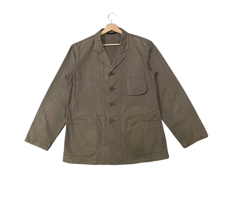 May include: A khaki-colored jacket with a button-up front, two large front pockets, and a single chest pocket. The jacket has long sleeves and a classic collar. The garment is hanging on a wooden hanger against a white background.