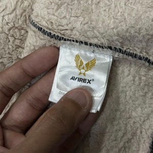 May include: A close-up of an Avirex label, featuring a gold emblem of wings and a star. The white label with black text is sewn into a beige, textured fabric. The label is held by a hand.