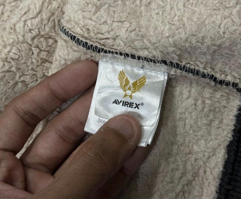 May include: A close-up of an Avirex label, featuring a gold emblem of wings and a star. The white label with black text is sewn into a beige, textured fabric. The label is held by a hand.
