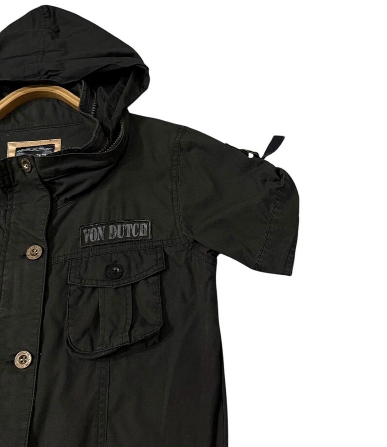 Puede incluir: Chaqueta negra con capucha con el texto "Von Dutch" en el pecho. La chaqueta tiene dos bolsillos delanteros, cierres de bot&oacute;n y una capucha con cord&oacute;n. Las mangas son cortas y tienen un detalle fruncido.
