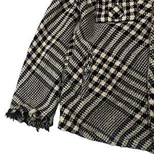May include: Close-up of a black and white houndstooth patterned jacket. The jacket has a classic design with a frayed edge on the sleeve. The fabric is a woven material with a repeating pattern.