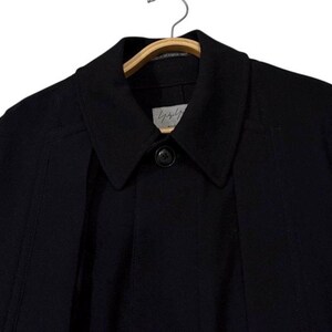 May include: A black coat with a collar and a single button closure. The coat is hanging on a wooden hanger. The brand name is visible on a small tag near the collar.