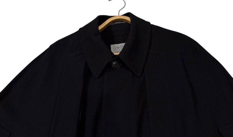 May include: A black coat with a collar and a single button closure. The coat is hanging on a wooden hanger. The brand name is visible on a small tag near the collar.