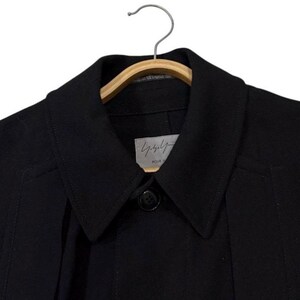 May include: Black coat with a notched collar and a single-button closure. The coat is hanging on a wooden hanger. A small label with Japanese text is visible near the collar.