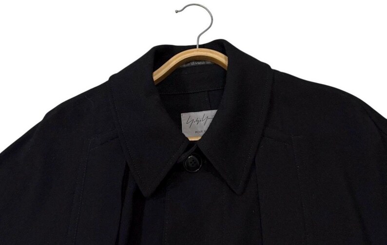 May include: Black coat with a notched collar and a single-button closure. The coat is hanging on a wooden hanger. A small label with Japanese text is visible near the collar.