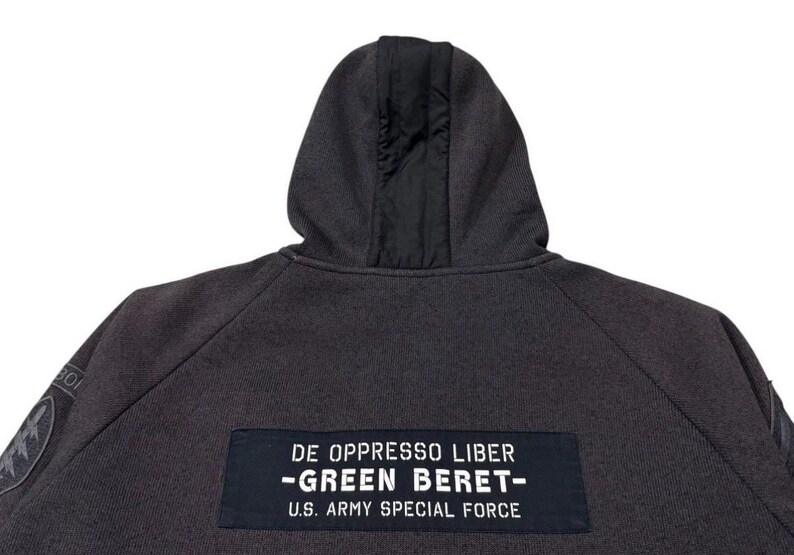 May include: Dark grey hoodie with a black hood lining. The back features a patch with the text "DE OPPRESSO LIBER - GREEN BERET - U.S. ARMY SPECIAL FORCE." Patches are visible on the sleeves. A casual, military-inspired garment.