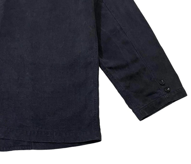 May include: A close-up shot of a navy blue jacket. The jacket features a buttoned cuff on the sleeve. The fabric looks to be a robust material, possibly denim or canvas. The jacket is a plain colour, with no visible patterns or designs.