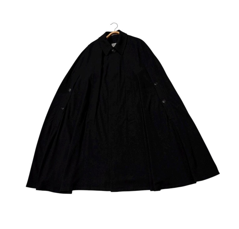 May include: A black cape-style coat with a button-down front and a collar. The coat is hanging on a wooden hanger. The fabric appears to be a medium-weight material, suitable for cooler weather. The coat has a classic, timeless design.