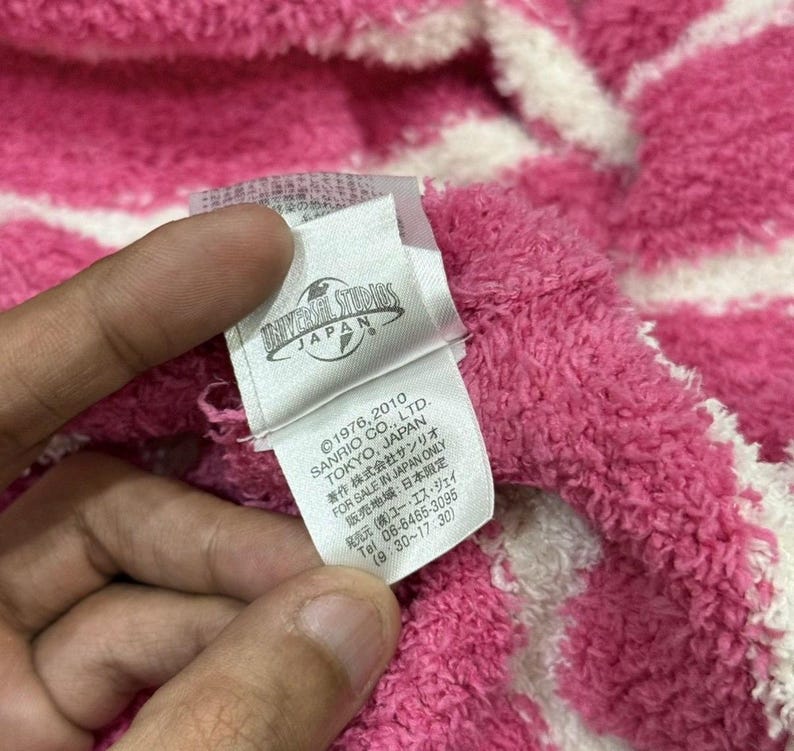 May include: Close-up of a pink and white striped fabric with a tag. The tag reads "UNIVERSAL STUDIOS JAPAN" and includes text about Sanrio Co., Ltd. and copyright information.