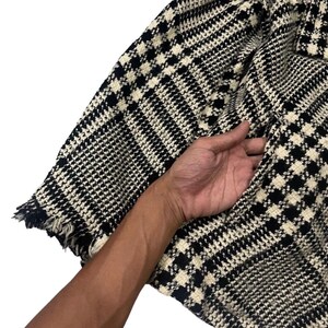 May include: Close-up of a black and white houndstooth patterned jacket. The jacket has a pocket and a frayed edge. The fabric is a woven material.