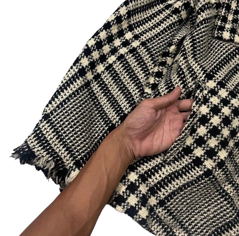 May include: Close-up of a black and white houndstooth patterned jacket. The jacket has a pocket and a frayed edge. The fabric is a woven material.