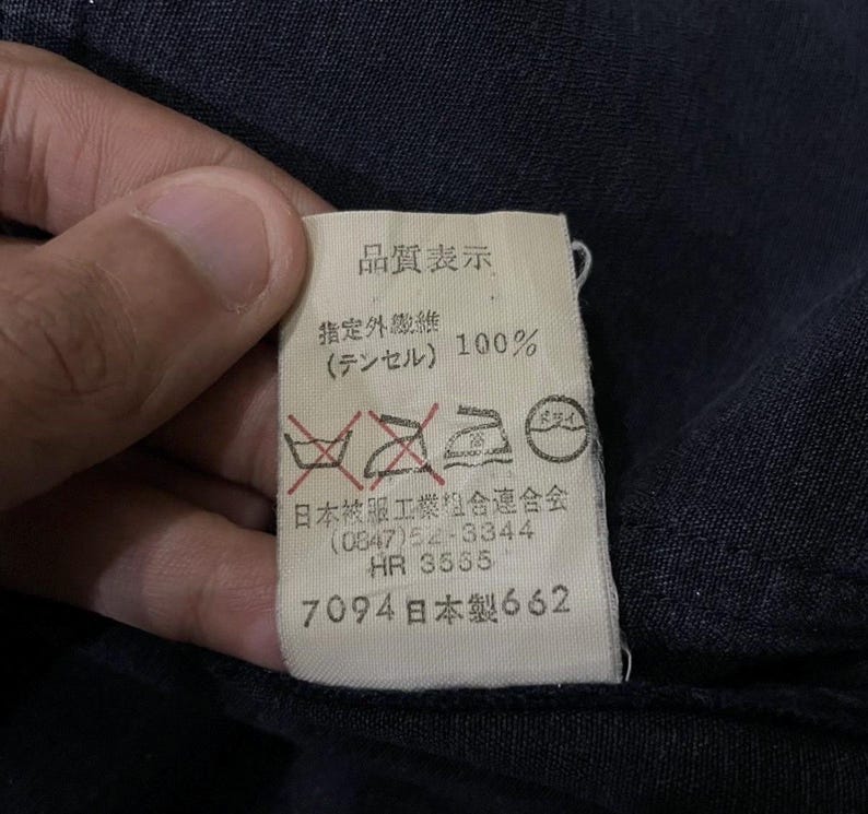 May include: A white label with Japanese text and symbols, including "品質表示" and "100%" in black. The label also features care symbols and the numbers "7094" and "662". The label is against a dark blue fabric.