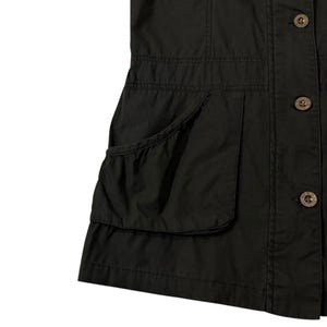 Puede incluir: Chaleco negro sin mangas con cierre de botones y dos bolsillos grandes. El chaleco tiene un corte entallado con costuras decorativas y botones marrones. La tela parece ser un material tejido duradero.