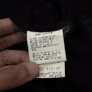 May include: A white tag with Japanese and English text, held against a black fabric. The tag includes care instructions about the material's nature, potential for pilling, and handling with care. The tag also has a product code.