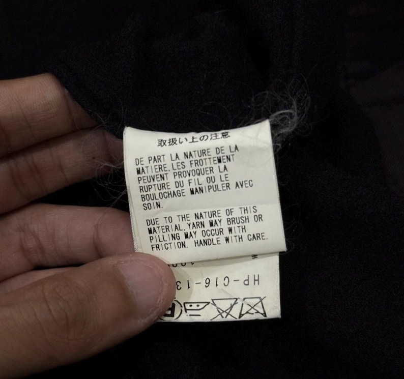 May include: A white tag with Japanese and English text, held against a black fabric. The tag includes care instructions about the material's nature, potential for pilling, and handling with care. The tag also has a product code.