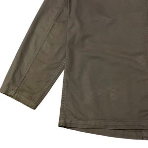 May include: Close-up of a muted olive green jacket. The jacket features a long sleeve and a button-down front. The fabric appears to be a durable cotton or blend, with visible stitching details along the seams. The overall style is casual and utilitarian.