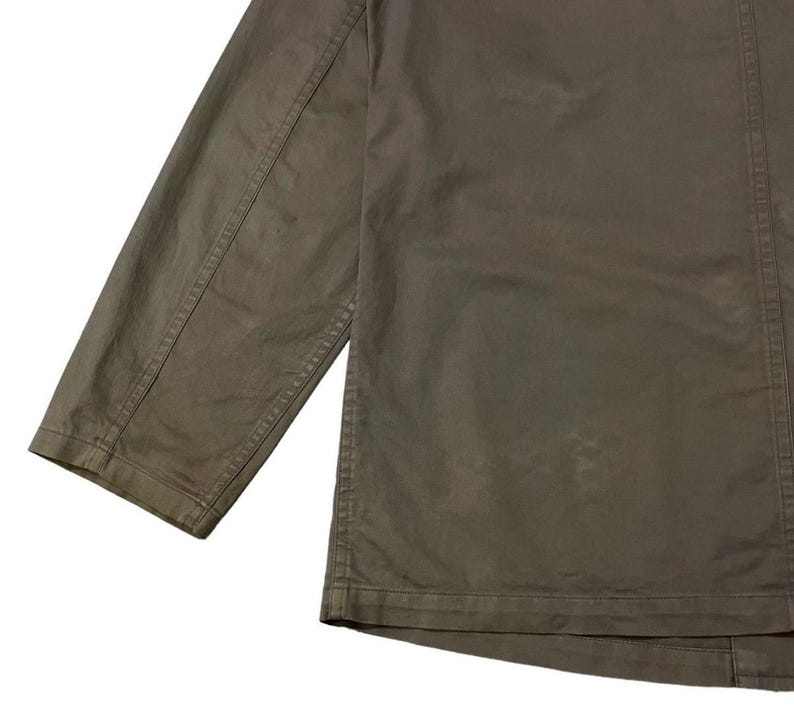 May include: Close-up of a muted olive green jacket. The jacket features a long sleeve and a button-down front. The fabric appears to be a durable cotton or blend, with visible stitching details along the seams. The overall style is casual and utilitarian.