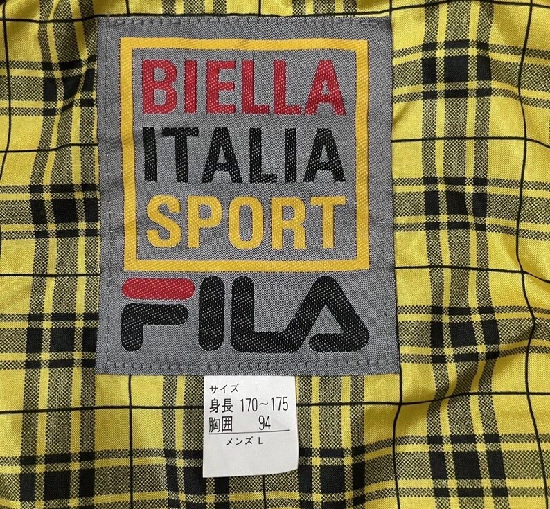 以下が含まれることがあります： 黄色と黒のチェック柄の生地のクローズアップ。ラベルには「BIELLA ITALIA SPORT」と赤、黒、黄色で書かれています。ブランド「FILA」も表示されています。ラベルはグレーで、黄色の縁取りがあります。