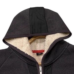 May include: A dark grey hooded sweatshirt with a full zip. The hood features a black panel down the centre and a cream-coloured, fuzzy lining. The sweatshirt is hanging on a wooden hanger.