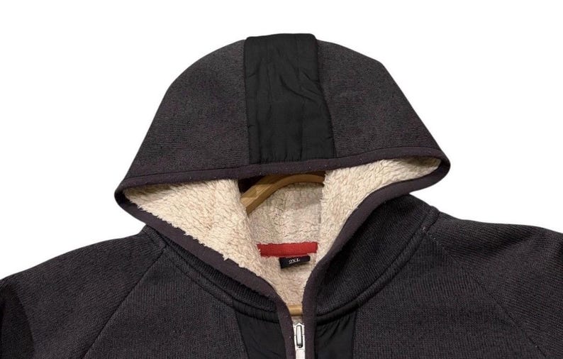 May include: A dark grey hooded sweatshirt with a full zip. The hood features a black panel down the centre and a cream-coloured, fuzzy lining. The sweatshirt is hanging on a wooden hanger.