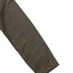 May include: Close-up of a khaki-colored jacket sleeve. The sleeve is angled across the frame, showcasing the fabric's texture and the stitching details along the seams. The jacket appears to be made of a durable material.