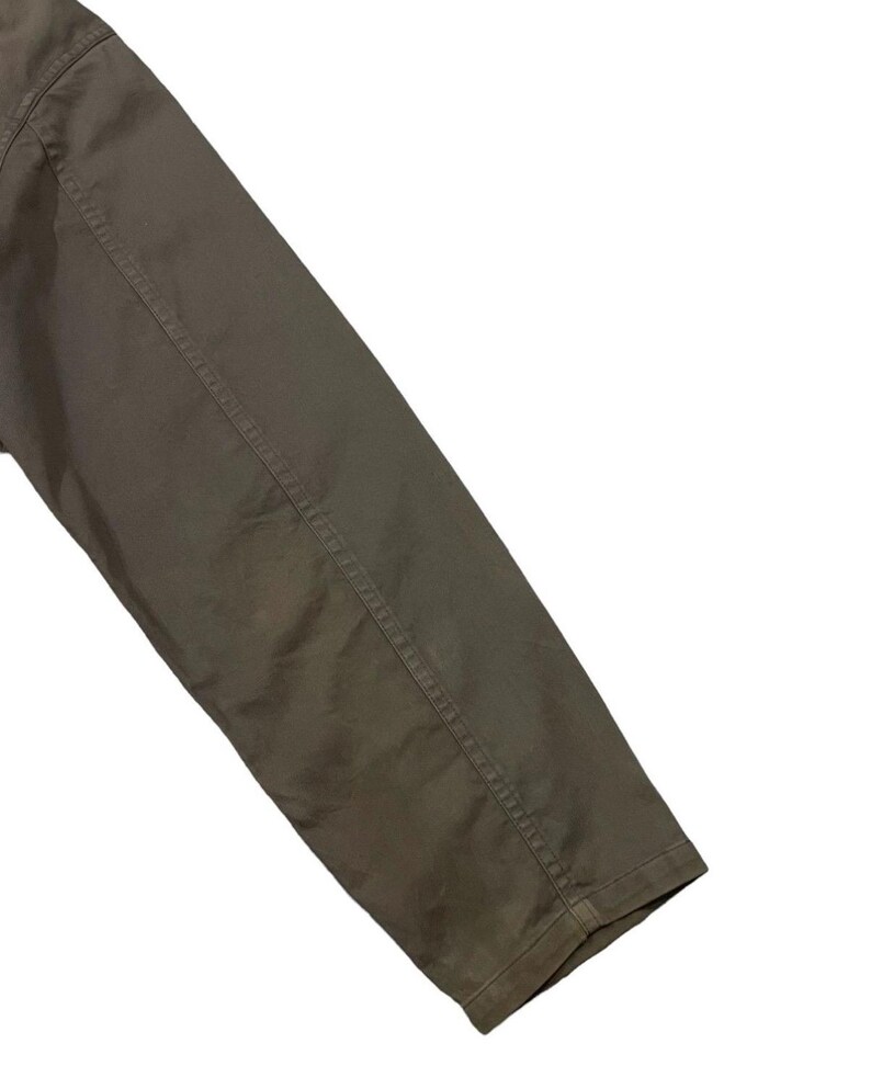 May include: Close-up of a khaki-colored jacket sleeve. The sleeve is angled across the frame, showcasing the fabric's texture and the stitching details along the seams. The jacket appears to be made of a durable material.