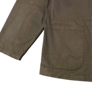 May include: Close-up of a khaki-colored jacket with three buttons and two front pockets. The jacket has a relaxed fit with three-quarter sleeves. The fabric appears to be a durable cotton or blend, suitable for casual wear. The jacket is a solid color.