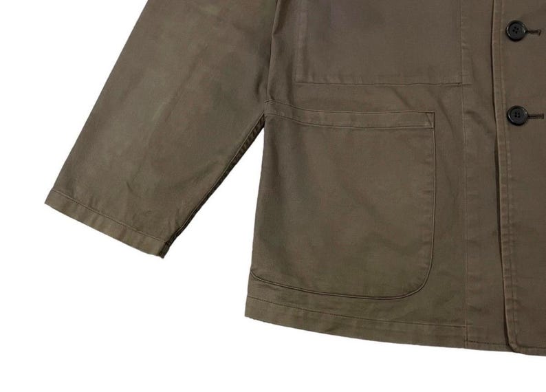 May include: Close-up of a khaki-colored jacket with three buttons and two front pockets. The jacket has a relaxed fit with three-quarter sleeves. The fabric appears to be a durable cotton or blend, suitable for casual wear. The jacket is a solid color.