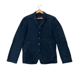 Puede incluir: Un blazer azul marino con cierre de botones y cuello con solapas. La chaqueta presenta detalles de costura blancos y dos bolsillos delanteros. Cuelga de una percha de madera.