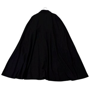 May include: A black cape with a buttoned collar, hanging from a hook. The cape is made of a dark fabric and has a wide, flowing design. The back view of the cape is shown, with the collar and shoulders visible.
