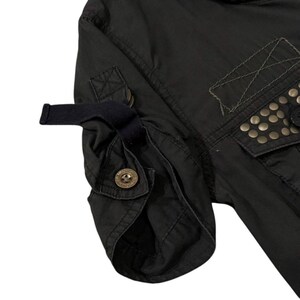 Puede incluir: Primer plano de una manga de chaqueta negra con pu&ntilde;o abotonado y detalle de correa. La chaqueta presenta un bolsillo con tachuelas met&aacute;licas y costuras decorativas. La tela parece ser un material oscuro y duradero.
