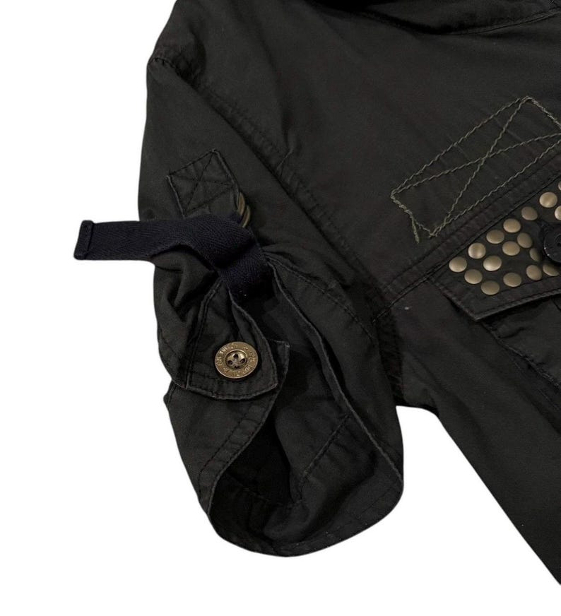 Puede incluir: Primer plano de una manga de chaqueta negra con pu&ntilde;o abotonado y detalle de correa. La chaqueta presenta un bolsillo con tachuelas met&aacute;licas y costuras decorativas. La tela parece ser un material oscuro y duradero.