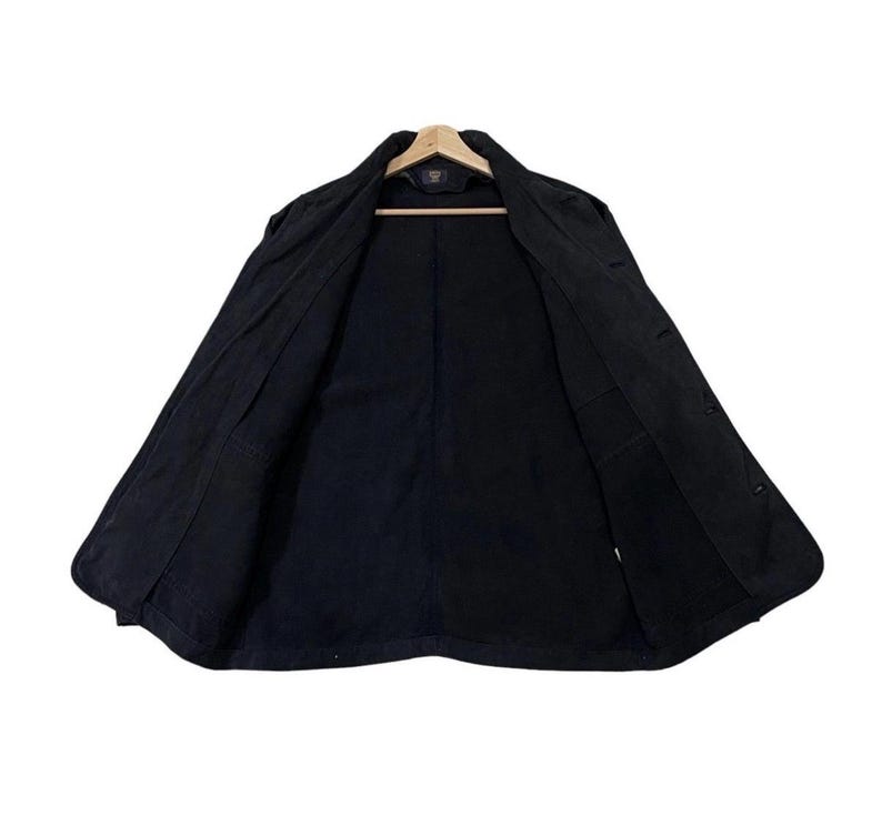 May include: A black jacket, open to show the interior, hanging on a wooden hanger. The jacket has a button-up front, a collar, and pockets. The lining is also black. The jacket appears to be made of a durable fabric.