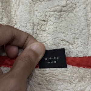 May include: A black label with the words "ESTABLISHED IN 1975" in white. The label is set against a cream-coloured, textured fabric with a red stripe.