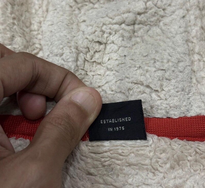 May include: A black label with the words "ESTABLISHED IN 1975" in white. The label is set against a cream-coloured, textured fabric with a red stripe.