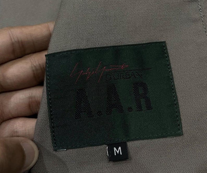 May include: Close-up of a dark green label with the text "Yohji Urban A.A.R." in black and red. The label is sewn onto a gray fabric. The label also has a size "M" tag.