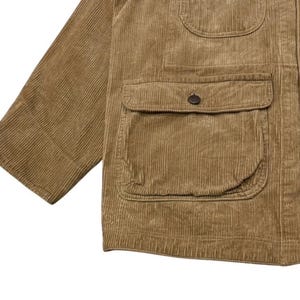 K&ouml;nnte beinhalten: Nahaufnahme einer beigefarbenen Cordjacke mit mehreren Taschen und Knopfverschl&uuml;ssen. Die Jacke hat ein klassisches Design mit einem strukturierten, gerippten Stoff. Der &Auml;rmel ist auf der linken Seite sichtbar.