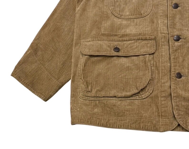 K&ouml;nnte beinhalten: Nahaufnahme einer beigefarbenen Cordjacke mit mehreren Taschen und Knopfverschl&uuml;ssen. Die Jacke hat ein klassisches Design mit einem strukturierten, gerippten Stoff. Der &Auml;rmel ist auf der linken Seite sichtbar.