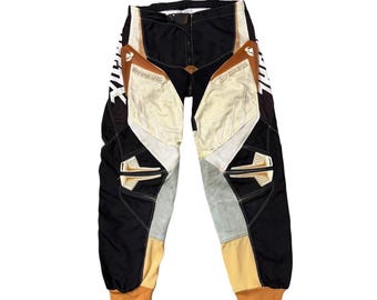 May include: Black, tan, and white motocross pants with reinforced panels and the text "The Motocross" printed on the sides. The pants have a padded design and are made of durable materials.