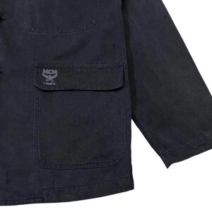 May include: A dark navy blue jacket with a button-up front, a chest pocket, and a sleeve. The jacket features the MCM logo embroidered on the pocket. The jacket is made of a durable fabric.