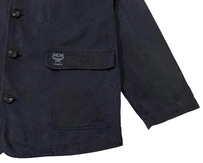 May include: A dark navy blue jacket with a button-up front, a chest pocket, and a sleeve. The jacket features the MCM logo embroidered on the pocket. The jacket is made of a durable fabric.