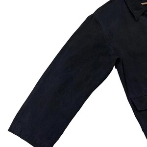 May include: A close-up of a dark navy blue jacket. The jacket has a collar, long sleeves, and a pocket. The fabric appears to be a sturdy material, possibly denim or canvas. The jacket is hanging on a wooden hanger.